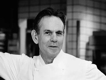 In a chef's coat, stands confidently in a professional kitchen with his arm resting on a tiled wall.