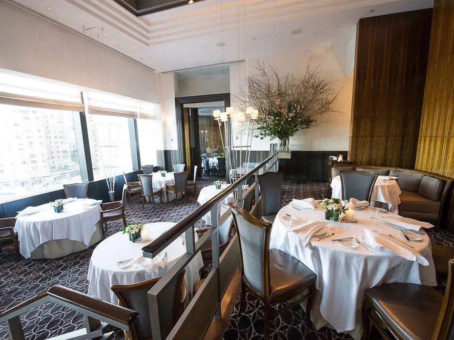 An upscale dining room with white-tablecloth tables, gray chairs, wood paneling, and large windows overlooking a city.
