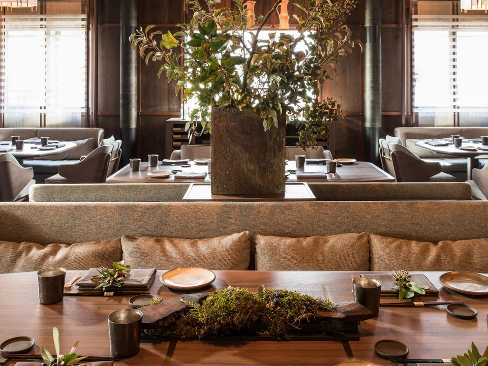 An elegant dining room with dark wood paneling, light-colored banquettes, and individual place settings with ceramic plates and metal cups. A large,