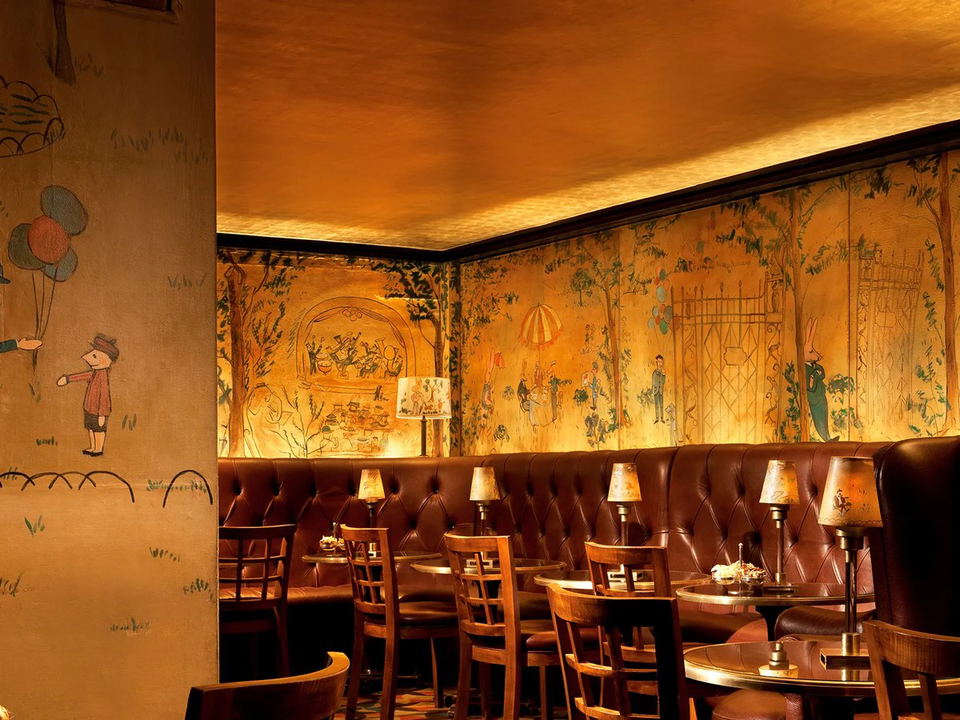 An intimate, warmly lit bar interior with hand-painted Ludwig Bemelmans murals depicting whimsical park scenes, tufted dark-leather banquettes, and