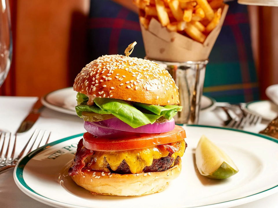 A cheeseburger with bacon, tomato, red onion, lettuce, and sesame bun, served with a lime wedge and fries in a metal cup on a white plate with a