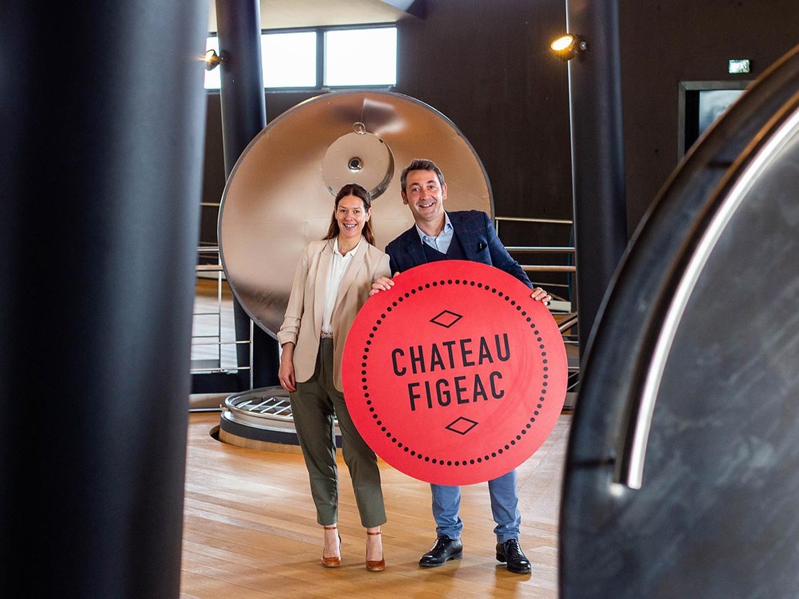 A man and a woman hold a large red circular sign with "CHATEAU FIGEAC" written on it, inside a modern winery.