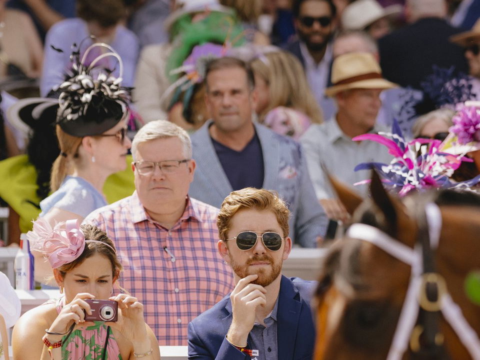 Well-dressed guests in fascinators and blazers watch horses at Churchill Downs Derby Day.