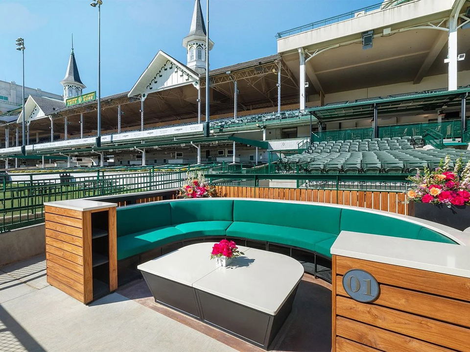 An outdoor seating pod with a curved teal sofa and a white coffee table, numbered "01", overlooks the Churchill Downs racetrack and grandstand with