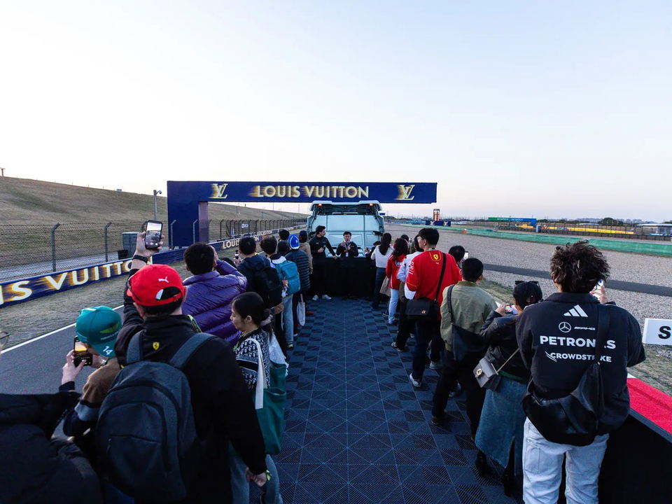 A crowd of F1 fans stands on a pit lane walkway under a blue Louis Vuitton arch, with a white truck and staff in the background.