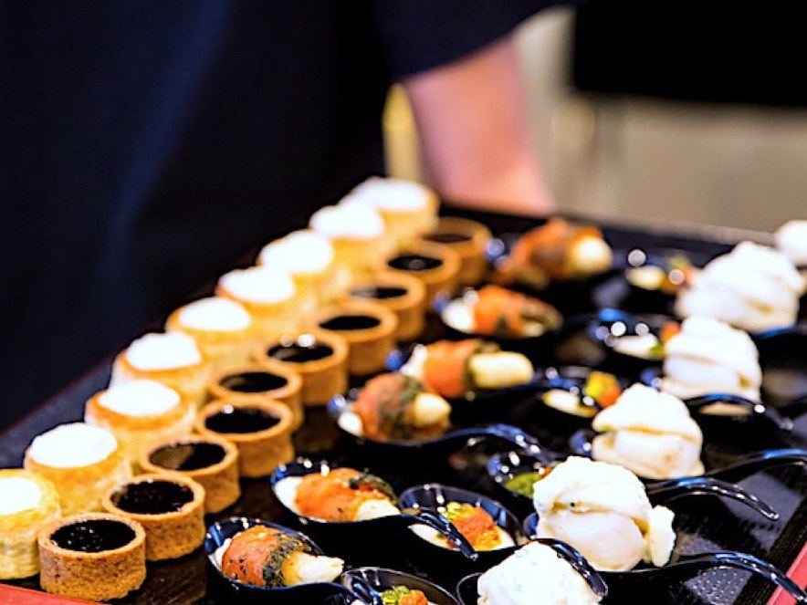 A server holds a black tray laden with an assortment of elegant canapés, including pastry tartlets with dark fillings, smoked salmon bites, and