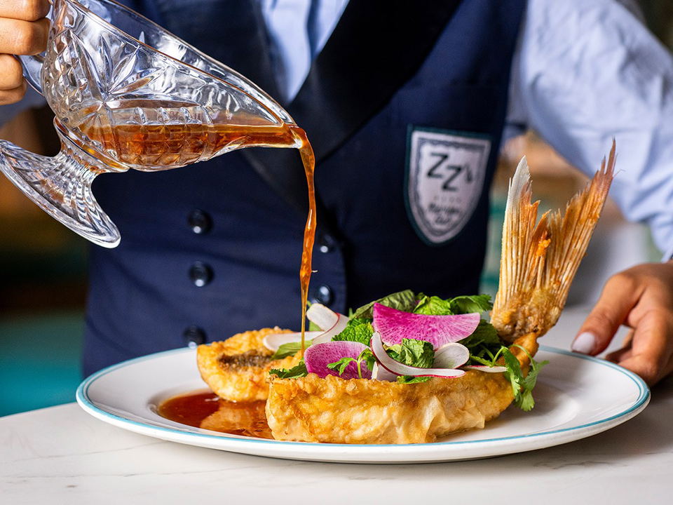 A server in a navy ZZ's Club uniform pours amber sauce from a crystal pitcher over a whole crispy snapper dish garnished with watermelon radish,