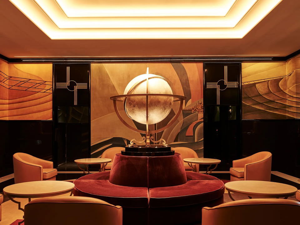 An opulent Art Deco lounge interior with a central illuminated globe sculpture, circular velvet banquette, peach armchairs, and amber murals.