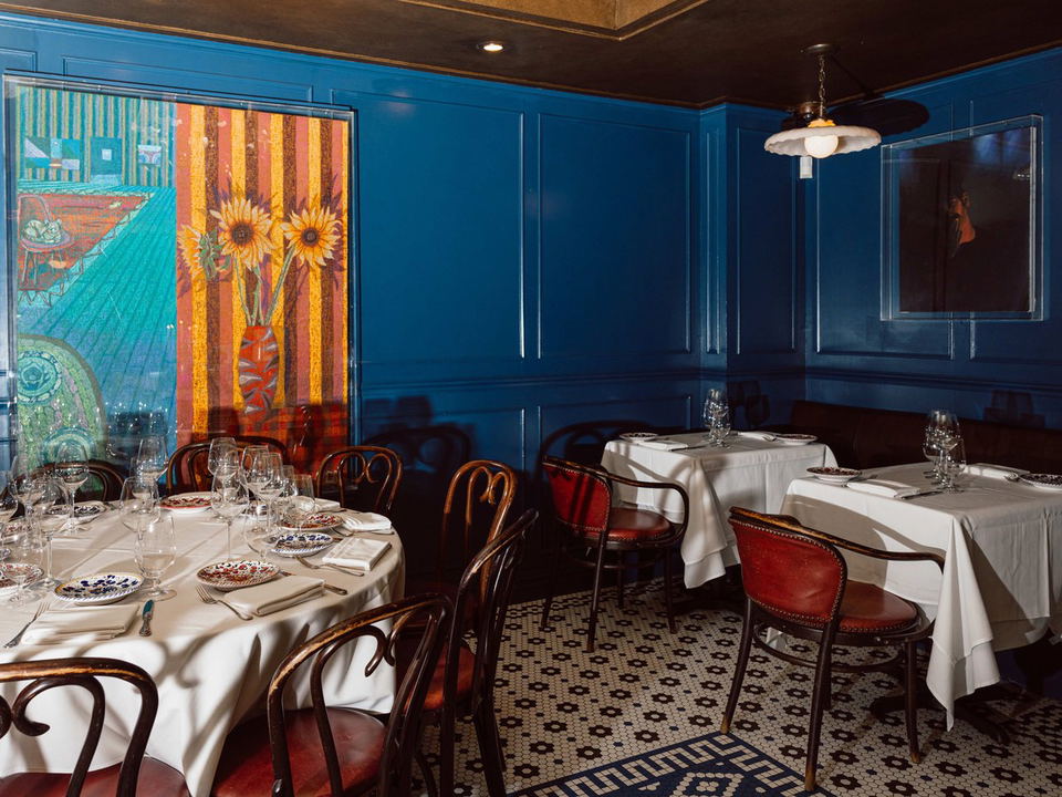 A dining room with blue paneled walls, white tablecloths, bentwood chairs, and a large, colorful painting of sunflowers.