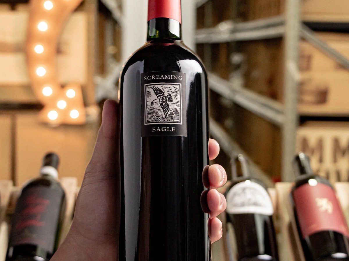 A hand holds a dark bottle of Screaming Eagle wine, featuring an eagle illustration on its label, in a dimly lit wine cellar.