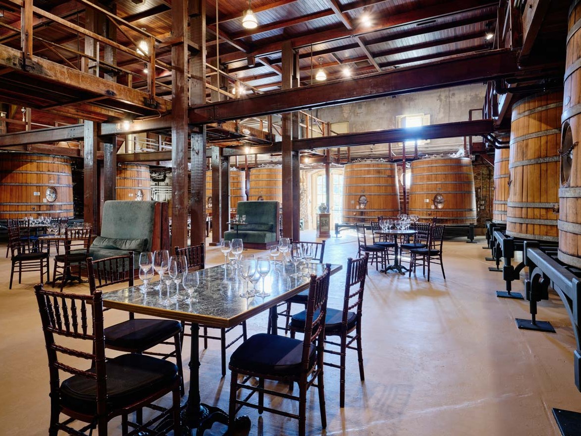 A large, rustic winery barrel hall with high ceilings and exposed wooden beams. Several massive wooden fermentation vats line the walls.