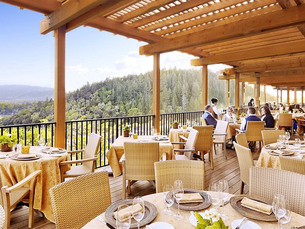 An upscale open-air terrace restaurant with wicker chairs, gold-toned tablecloths, set tables with wine glasses, and sweeping valley views.