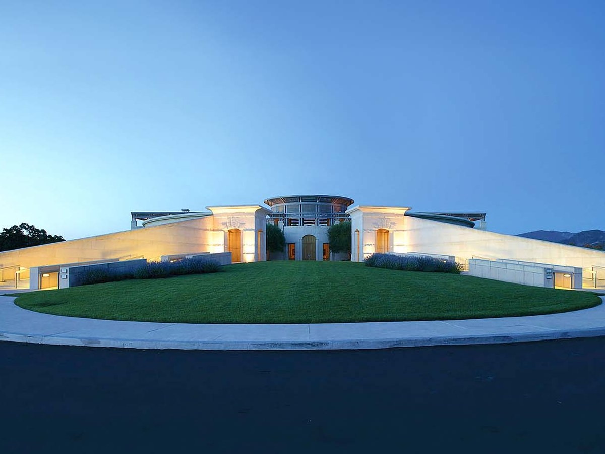 A modernist architecture at dusk, featuring a curved roofline, illuminated forecourt, and a circular grass roundabout.