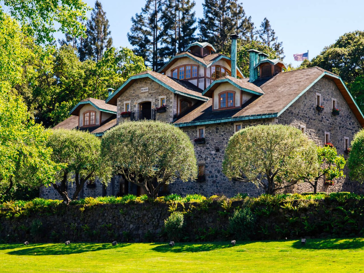 A historic stone building with multiple dormer windows and copper-trimmed roofs, surrounded by trees and a green lawn with a pond.