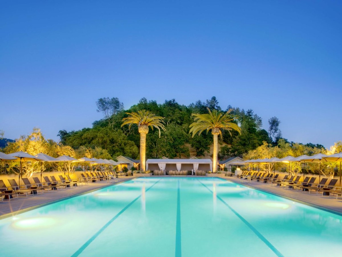 A resort pool at dusk, with lounge chairs, umbrellas, palm trees, and cabanas, backed by a wooded hillside.