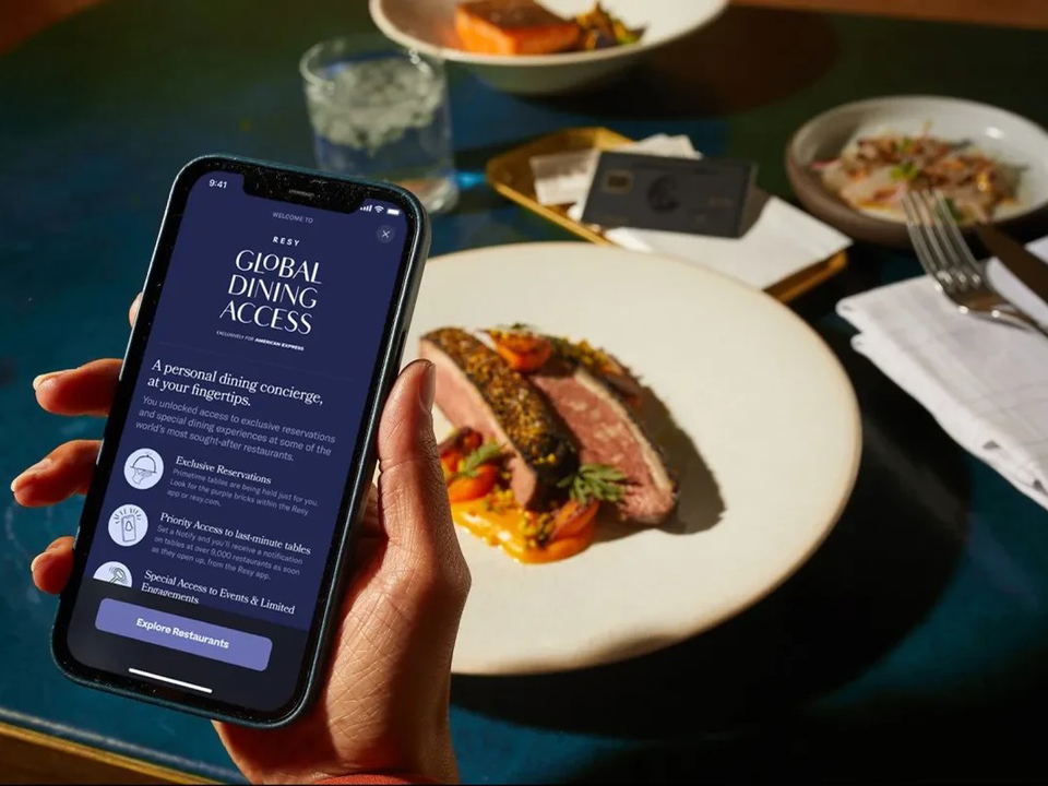 A hand holds a smartphone displaying the Resy Global Dining Access app screen in front of an upscale restaurant table with a plated meat dish.
