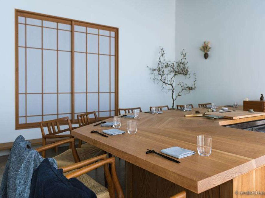 A minimalist Japanese-influenced dining room with a long, light wood counter table set with chopsticks, napkins, and water glasses.