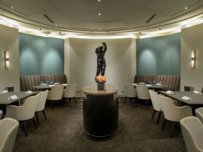 An elegant fine-dining room with curved teal walls, a central pedestal with a dark sculpture and roses, and upholstered banquettes.