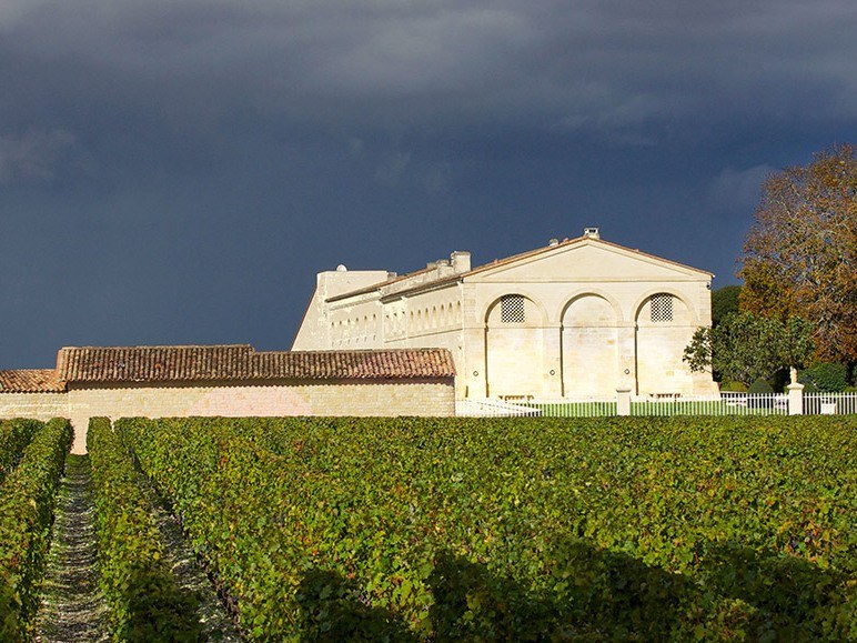 White stone winery building with arched windows and terracotta roof, surrounded by green vineyard hedges under dramatic sky.
