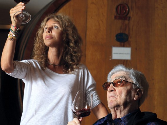 An older man in a wheelchair and a younger woman examine glasses of red wine in a cellar with large wooden barrels.