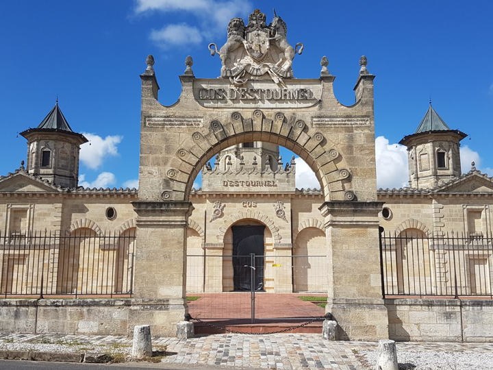 The ornate stone entrance gate of Cos d'Estournel, featuring a central archway with 'COS D'ESTOURNEL' and 'DESTOURNEL' inscriptions, flanked by small