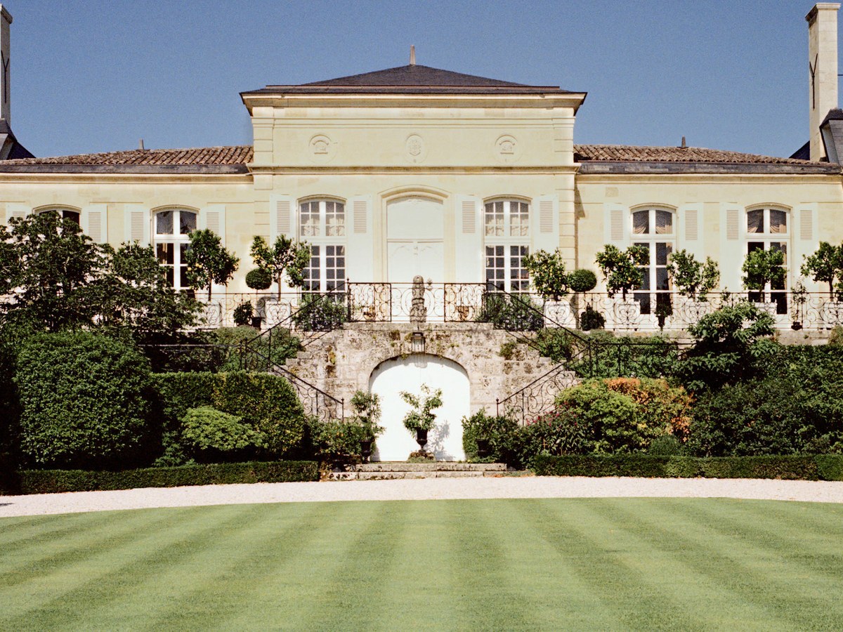 A grand, pale stone château with a dark mansard roof and dormer windows, featuring an ornate iron balustrade, manicured lawn, and formal garden