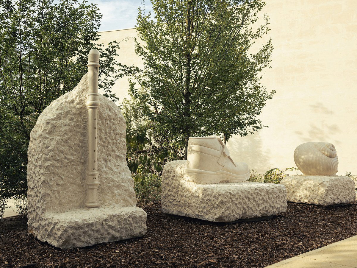 Three large, rough-hewn white stone sculptures: a tall flute, a work boot, and a snail shell, set among green trees.