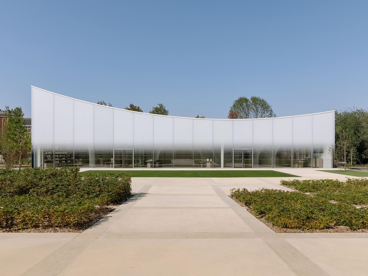 A modern, curved pavilion with translucent glass panels and white framing, surrounded by green hedges and trees under a clear blue sky.