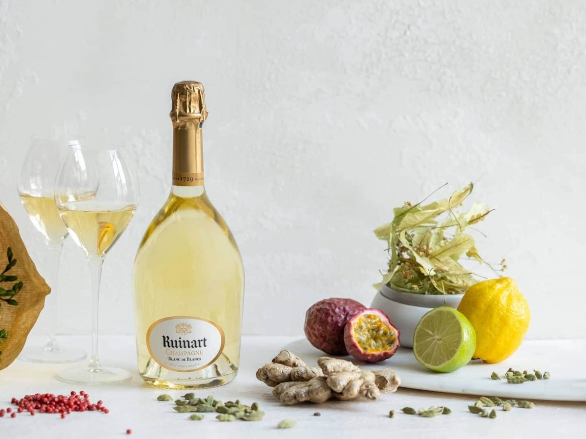 A bottle of Ruinart Blanc de Blancs Champagne, two glasses of wine, white flowers, and various fruits and spices.
