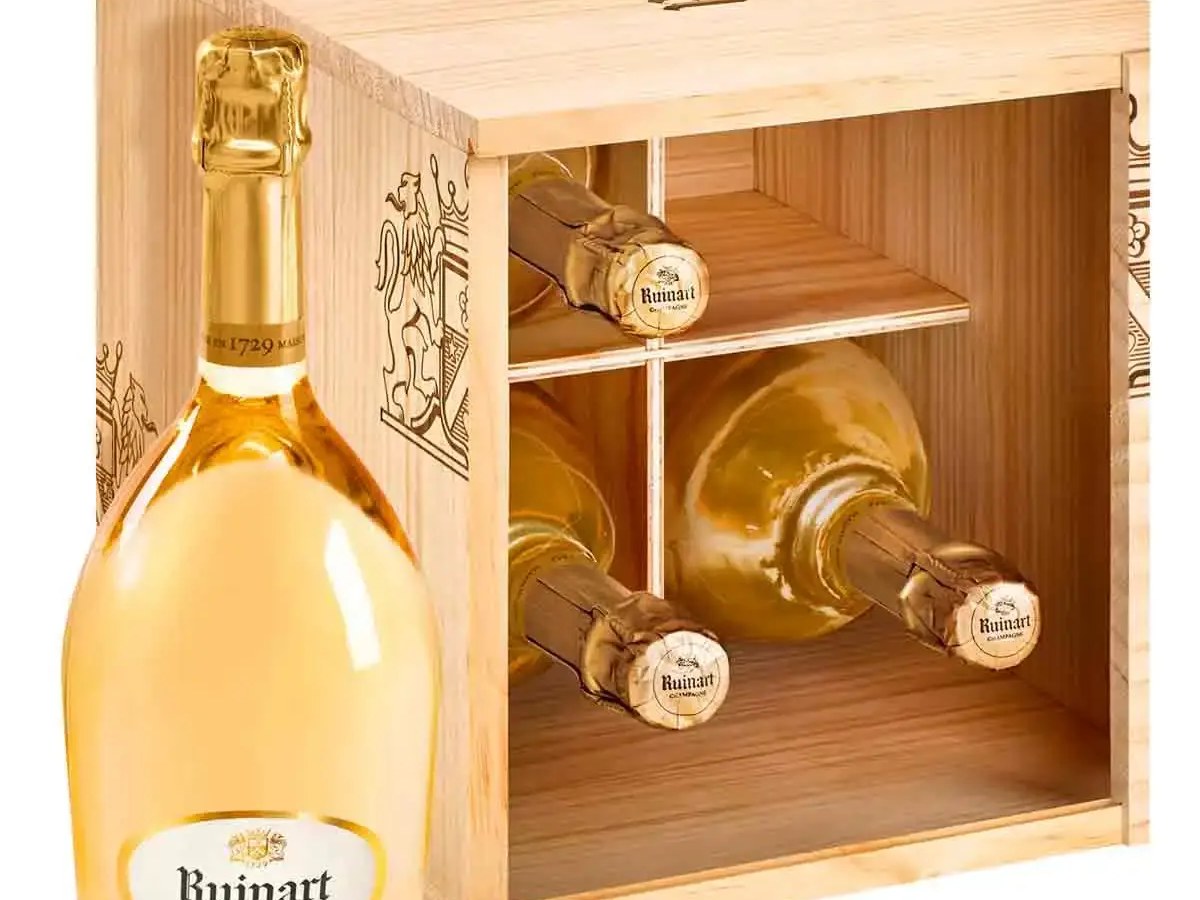 A bottle of Ruinart Blanc de Blancs Champagne stands next to a wooden cellar box holding three more bottles.