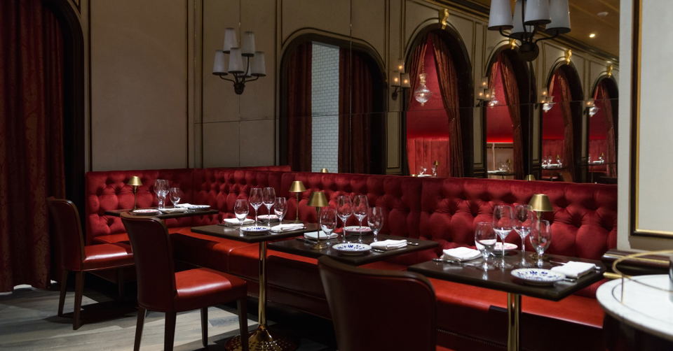 A luxurious restaurant interior with red velvet booth seating along the walls, black dining tables, brass lighting fixtures, and mirrored archways.