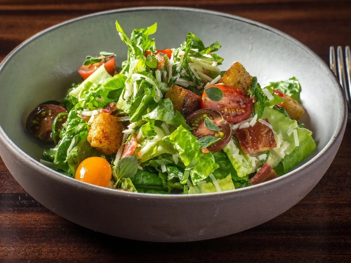 A close-up of the Canlis Salad in a light gray bowl, featuring fresh green lettuce, halved cherry tomatoes, golden croutons, and shaved cheese.