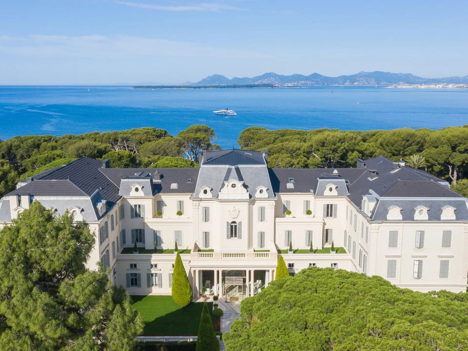 An aerial view of the grand white Belle Époque Hôtel du Cap-Eden-Roc, surrounded by green pine trees, with the blue sea and distant coastline visible
