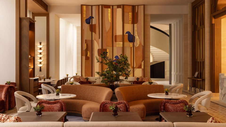 A modern luxury hotel lobby with curved furniture in earth tones, decorative wooden wall panels with blue bird motifs, potted plants, and marble-topped side tables.