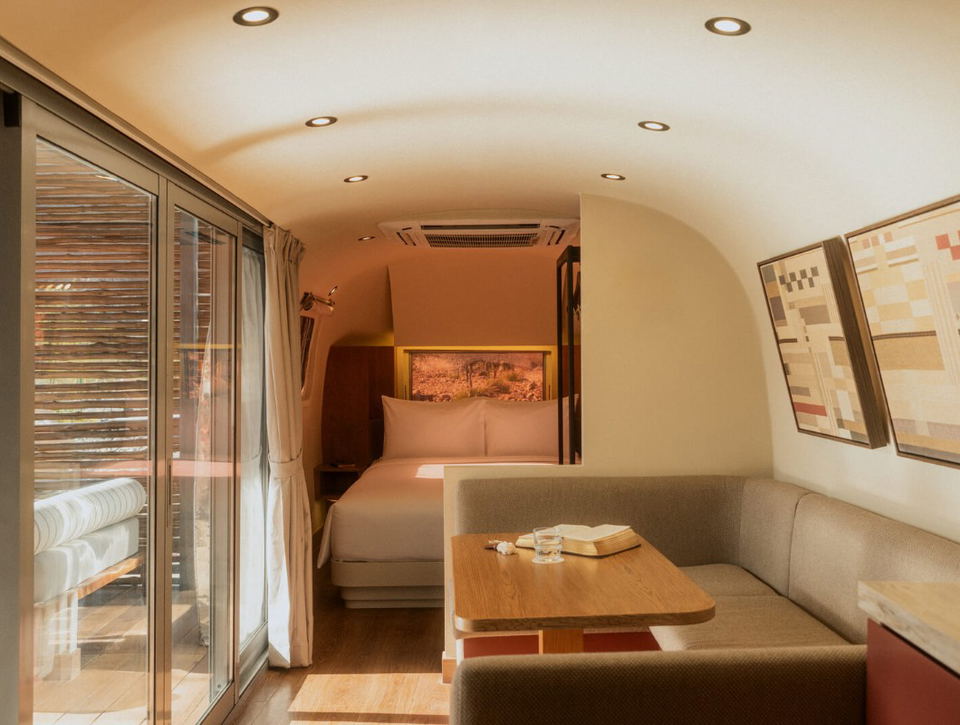Interior of a modern caravan/Airstream showing a queen bed with terracotta-toned headboard, neutral sand and beige tones, wooden table, gray sofa seat