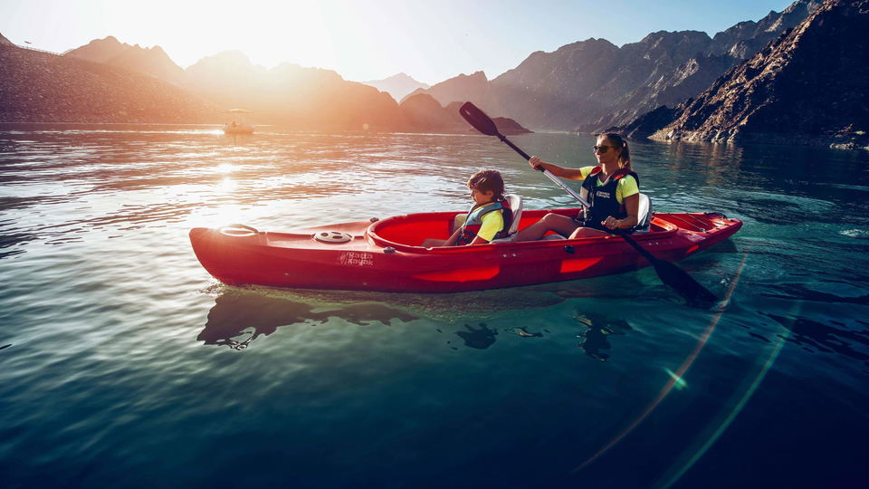 Two people kayaking in a red tandem kayak on clear turquoise water with dramatic mountain peaks (Hajar Mountains) in the background; the source page f