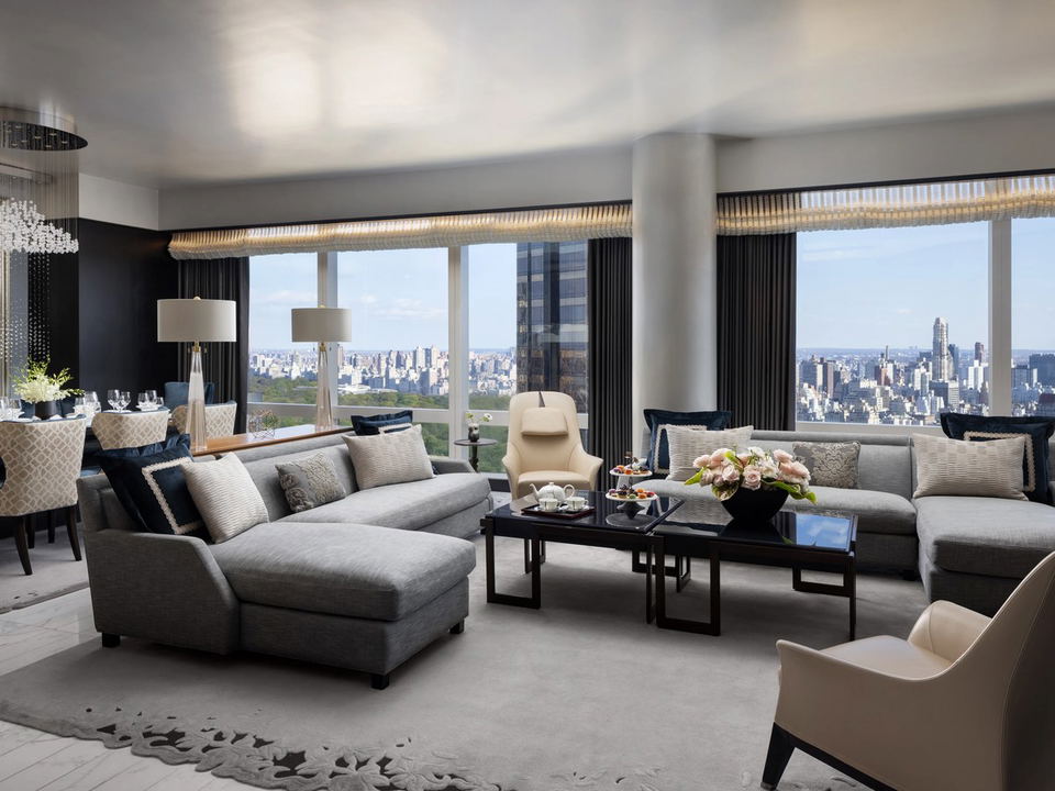 A luxurious hotel suite living room with floor-to-ceiling windows overlooking Central Park and the Manhattan skyline.