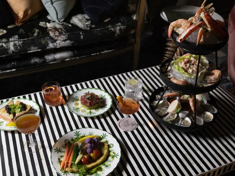 An overhead shot of a striped cocktail table set with a seafood tower, small plates, and cocktails in a dimly lit lounge interior.
