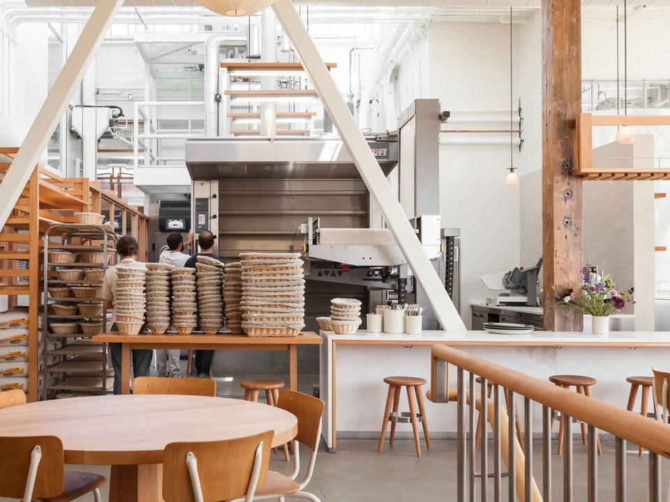 Interior of Tartine Manufactory in San Francisco, showing an open bakery with large commercial ovens, stacked proofing baskets, and café seating.
