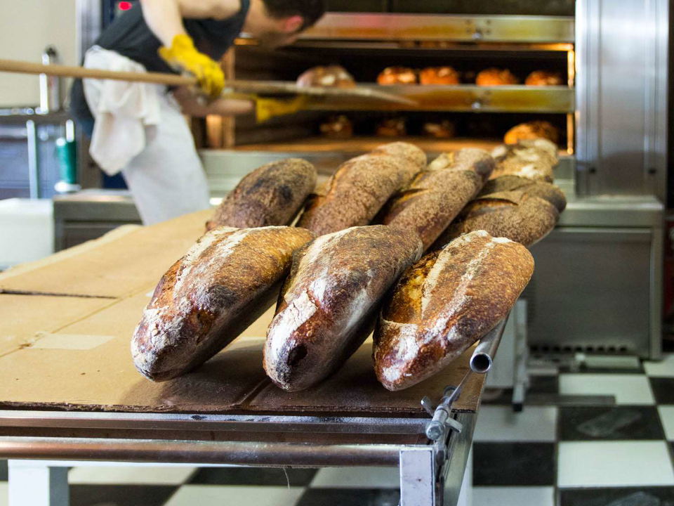 A baker in yellow gloves uses a long wooden peel to remove loaves of Tartine bread from a large commercial oven, with many freshly baked loaves