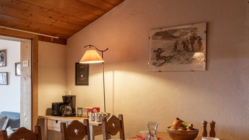 Interior of a rustic dining room with wooden ceiling beams, a table with wooden chairs, a floor lamp with amber shade, coffee maker, fruit bowl, and sepia-toned artwork on the walls.