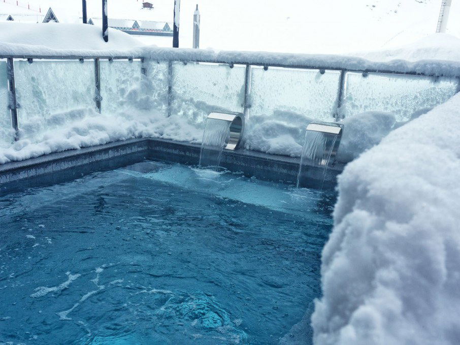 An outdoor heated swimming pool surrounded by snow, with two water features flowing into the pool, set against a backdrop of snowy mountains and