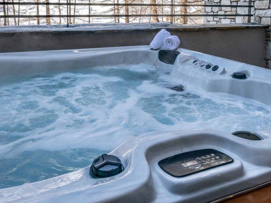 A steaming hot tub with multiple jets and digital controls on a snow-covered deck with white towels nearby and winter trees visible in the background.
