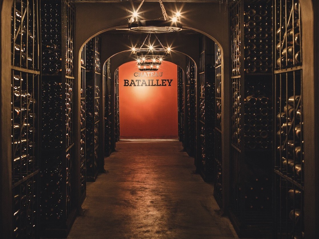 A long, dark wine cellar with arched ceilings and rows of wine bottles in metal racks, leading to a brightly lit red wall with
