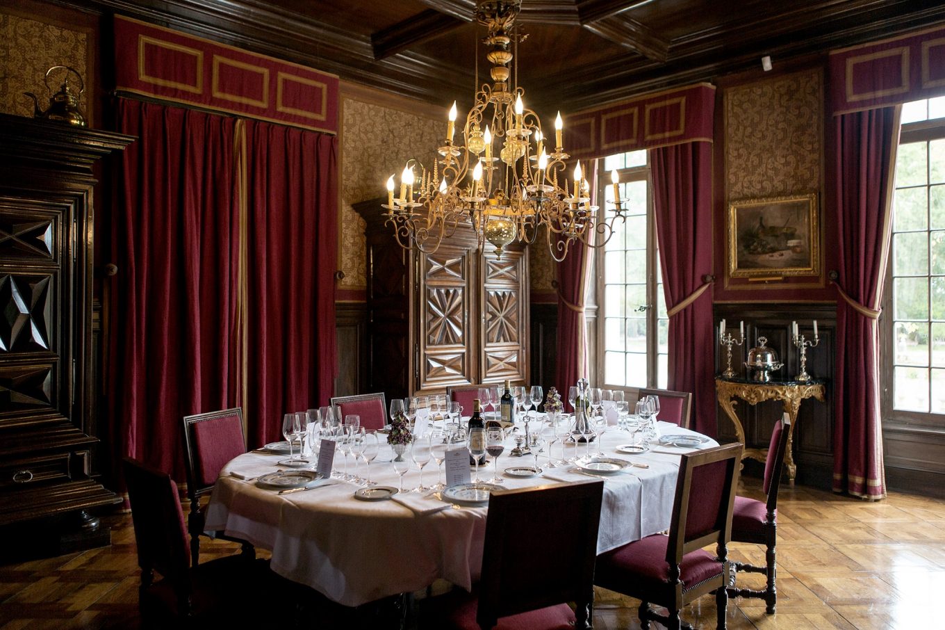 Ornate dining room at Château Batailley in Pauillac, with crimson curtains, carved wood paneling, a brass chandelier, and an oval table formally set with wine glasses and a bottle of Batailley red.