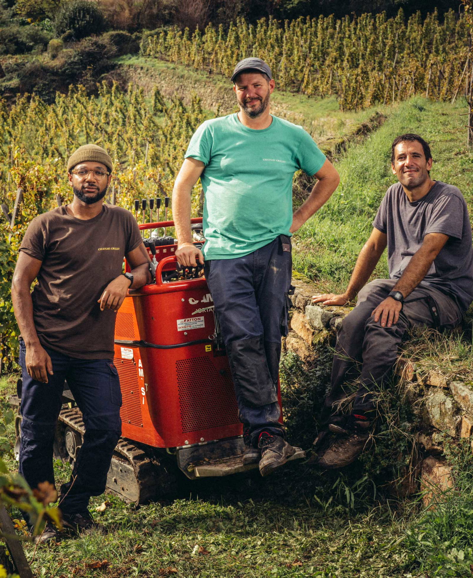 Three men with a red FATTON grape press in a vineyard; brown and green t-shirts visible
