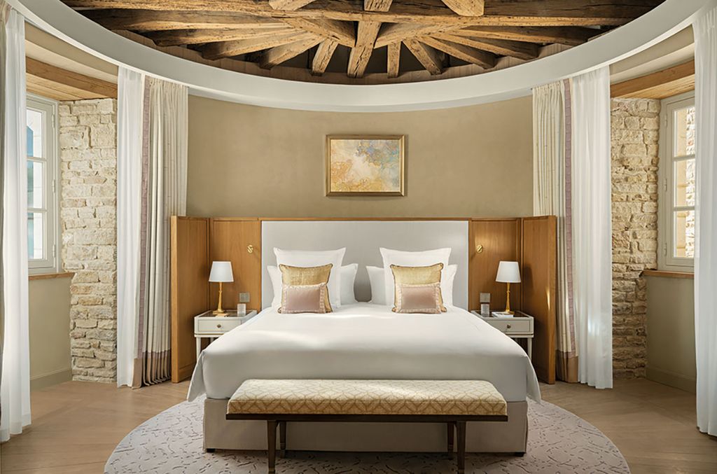 An elegant bedroom with a white upholstered bed, wooden nightstands with lamps, curved walls with stone accents, cream curtains, and decorative wooden ceiling beams.