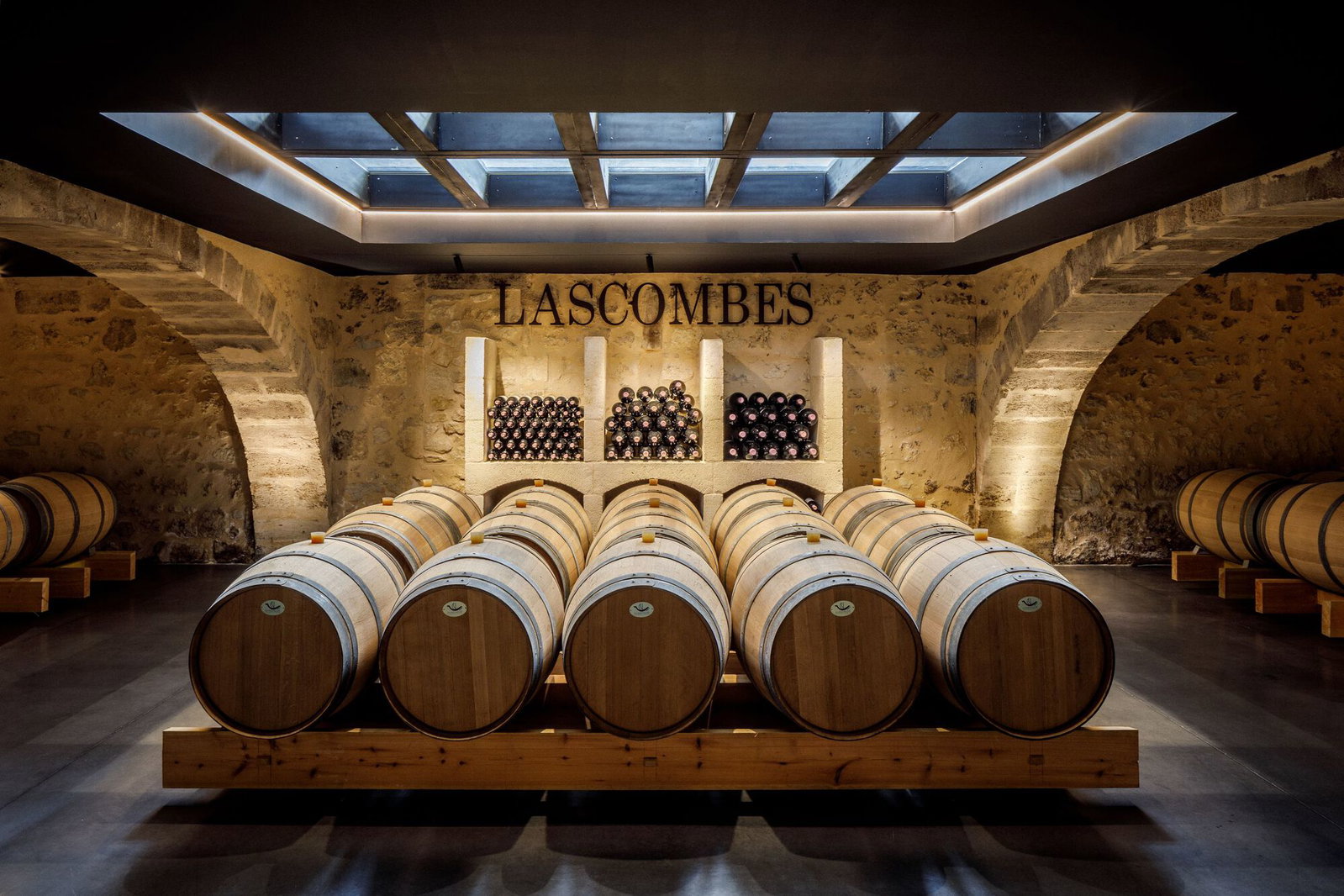 Rows of new oak barrels fill the stone-vaulted chai at Château Lascombes, with bottle alcoves and the estate name on the back wall beneath a glass skylight.