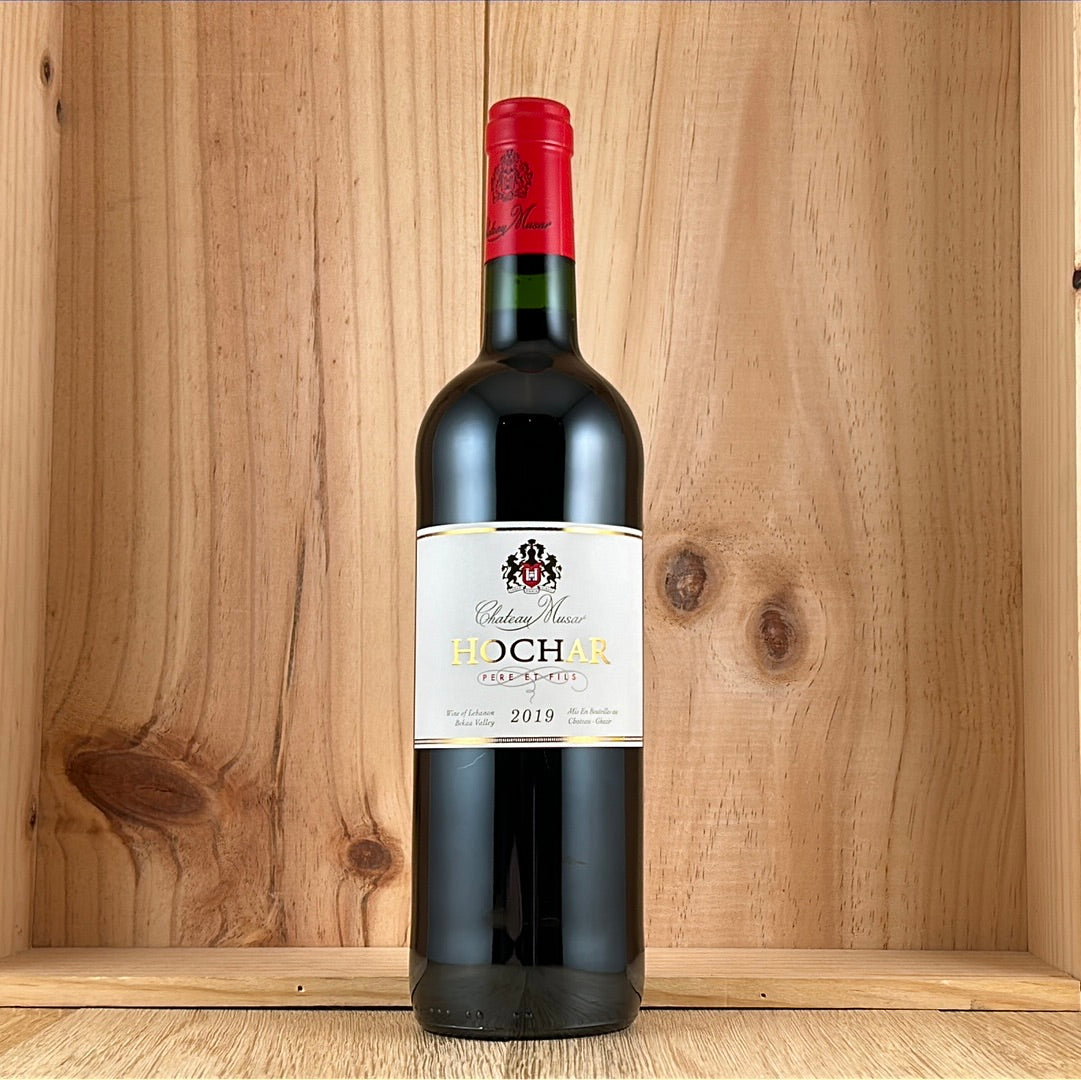 Bottle of Chateau Musar Hochar Pere et Fils 2019 red wine, with red capsule and white label, displayed against a wooden wine rack.
