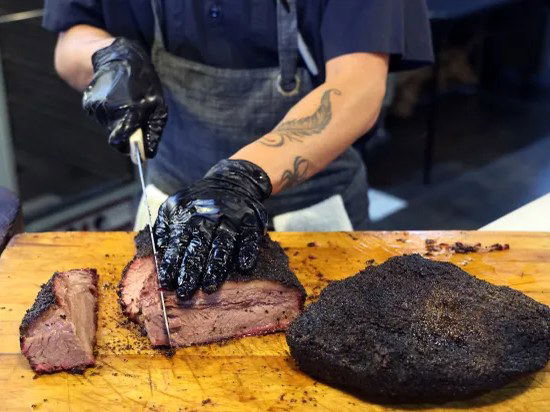 A person in black gloves and an apron slices a large piece of smoked brisket on a wooden cutting board, with another piece already cut.